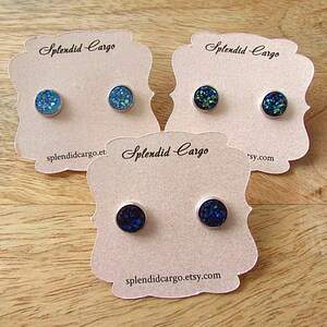 May include: Three pairs of stud earrings with sparkling, iridescent blue, teal, and navy blue surfaces. Each earring is set in a silver-colored metal. The earrings are displayed on pink cards with the text "Splendid Cargo" and a website address.