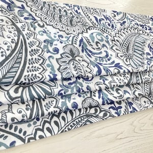 May include: Folded fabric with a white background and a repeating paisley pattern in shades of blue and gray. The fabric is layered, showcasing the intricate design of the floral and teardrop motifs. The pattern is detailed and visually appealing.