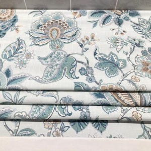 May include: A fabric roman shade featuring a repeating floral pattern in shades of teal, light blue, and muted brown on a white background. The textile displays large stylized blossoms, intricate leaves, and winding stems across the surface.