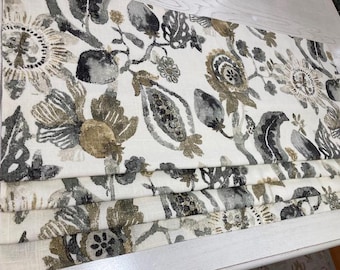 May include: A folded fabric with a botanical pattern in shades of gray, beige, and black. The design features stylized flowers, leaves, and seed pods on a cream-colored background. The fabric is layered, showing the pattern's repeat.