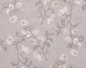 May include: A close-up of a fabric with an embroidered floral pattern. The design features white and light tan flowers with gray stems and leaves on a light purple background. The fabric has a textured appearance.