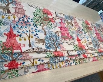 May include: A white fabric roman shade with a colorful pattern of pink and red pagodas, green trees, blue clouds, and brown leopards. The fabric has a pleated design.
