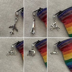May include: A selection of silver travel-themed charms: an aeroplane, rocket, motorcycle, caravan, and car. Each charm is attached to a black cord with a silver clasp. The background is a rainbow-coloured fabric.