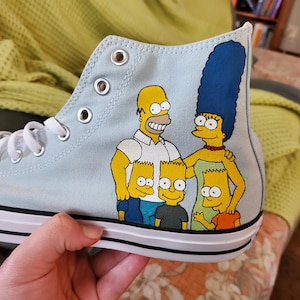 Puede incluir: Zapatillas altas de color azul claro con un diseño pintado a mano de la familia Simpson. Homero, Marge, Bart, Lisa y Maggie están representados con su piel amarilla icónica y su ropa característica.