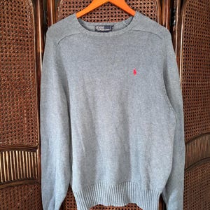 Könnte beinhalten: Ein grauer Rundhals-Pullover mit rotem Logo auf der Brust. Der Pullover hängt an einem Holzbügel. Der Pullover hat gerippte Bündchen und Saum. Der Markenname ist auf dem Etikett sichtbar.