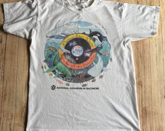 May include: White t-shirt featuring a circular graphic with marine life, including whales, sharks, and fish, surrounding a planet Earth image. The text reads "Preserve Their World" and "National Aquarium in Baltimore".