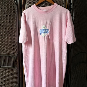 Könnte beinhalten: Hellrosa T-Shirt mit einem rautenförmigen Logo mit dem Schriftzug "Guess" in Blau. Das T-Shirt hängt an einem Holzbügel. Es hat einen Rundhalsausschnitt und kurze Ärmel. Dunkler Hintergrund.