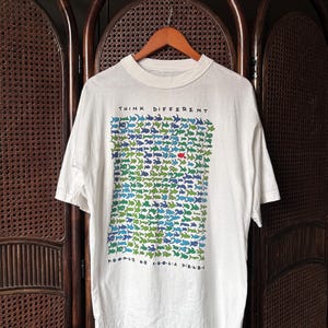 Könnte beinhalten: Weißes T-Shirt mit dem Schriftzug "THINK DIFFERENT" über einem Muster aus blauen, grünen und roten Fischen. Die Worte "PEOPLE OF POOLA FIELDS" sind darunter gedruckt. Das T-Shirt hängt an einem Holzbügel.