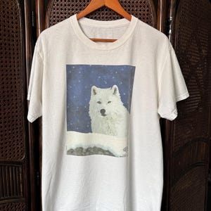 Pode incluir: Uma t-shirt branca de manga curta com uma impressão gráfica retangular no peito. A estampa retrata um lobo branco na neve contra um céu noturno azul escuro estrelado. A t-shirt tem gola redonda e está pendurada num cabide de madeira.