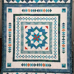 Puede incluir: Una colcha con un patrón geométrico azul, verde y blanco. El centro de la colcha presenta un diseño de estrella con acentos rojos y naranjas.