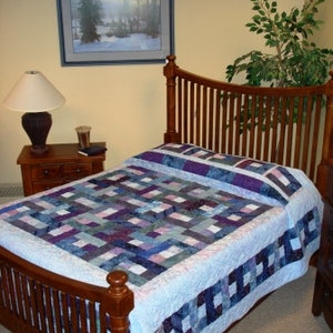 Op de afbeelding: Een houten bedframe met een hoofdbord en voetbord. Het bed is bedekt met een quilt in tinten blauw, paars en wit. Een nachtkastje met een lamp en een ingelijste foto zijn zichtbaar.