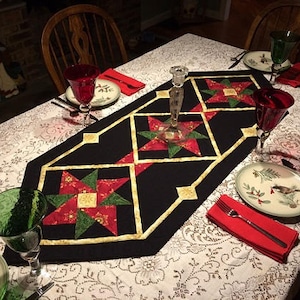 Może przedstawiać: Czarny bieżnik stołowy z geometrycznym wzorem czerwonych, zielonych i złotych gwiazd. Bieżnik leży na białym obrusie z koronkowym wzorem. Na stole stoją dwa czerwone kieliszki do wina, jeden zielony kieliszek do wina i talerz z wzorem ptaka.