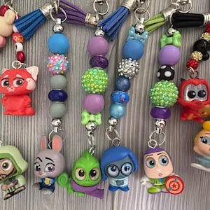 May include: A collection of colorful beaded keychains featuring various cartoon character charms. Each keychain has a unique combination of beads in shades of green, blue, purple, and pink, with silver-tone metal accents and a tassel.