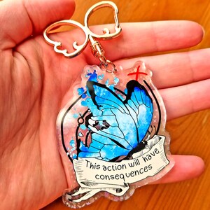 May include: A blue butterfly with watercolour wings is featured on a clear resin keychain. The keychain has a gold metal ring and a white ribbon with the text "This action will have consequences".