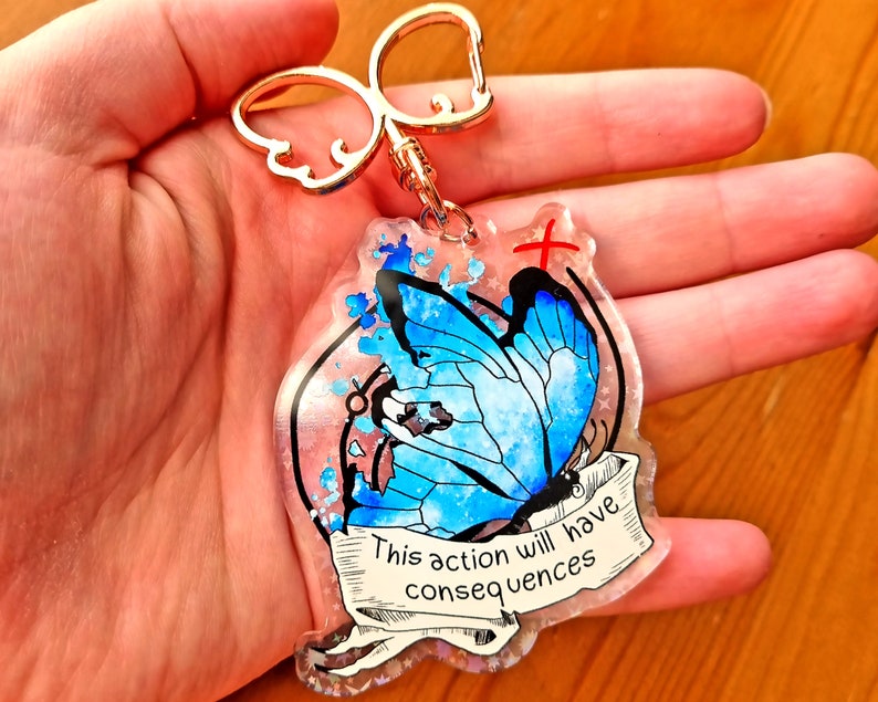May include: A blue butterfly with watercolour wings is featured on a clear resin keychain. The keychain has a gold metal ring and a white ribbon with the text "This action will have consequences".