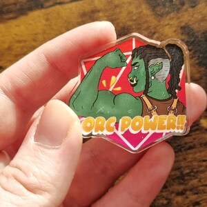 May include: A green orc with dreadlocks flexes their bicep in a pink and white background. The text "ORC POWER!" is written in yellow letters.