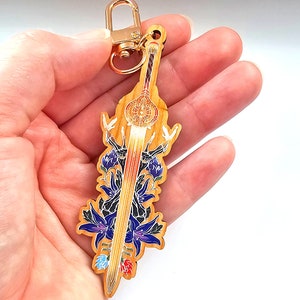 May include: A wooden keychain with a gold-coloured clasp, featuring a stylized sword design. The sword has a light brown wooden base with a central golden sunburst design and a red and gold blade. Blue and purple floral accents surround the sword.