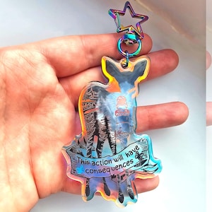May include: A holographic keychain featuring a black silhouette of a deer with a lighthouse in the background. The keychain has a rainbow iridescent finish and a silver star charm. The text "This action will have consequences" is printed on a banner below the deer.