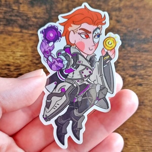 Pu&ograve; includere: Un adesivo fustellato raffigurante un personaggio stilizzato con capelli corti arancioni, che indossa un abbigliamento tattico bianco e grigio con accenti viola. Il personaggio ha un occhio rosso e una benda meccanica, tenendo un globo viola e una moneta d'oro.