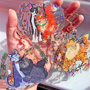 May include: Three clear acrylic keychains featuring colourful cat illustrations with holographic star accents. The designs include a black and white cat with an orange tabby, a blue and grey cat with a fluffy black cat, and two orange cats.