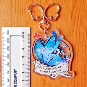 May include: A blue butterfly with glitter accents on a clear acrylic keyring. The keyring has a gold-coloured metal ring and a quote that reads "This action will have consequences".