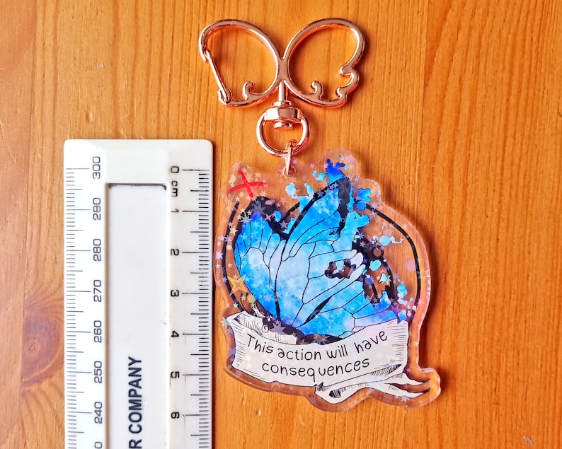 May include: A blue butterfly with glitter accents on a clear acrylic keyring. The keyring has a gold-coloured metal ring and a quote that reads "This action will have consequences".