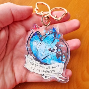 May include: A blue butterfly with glitter accents on a clear acrylic keyring. The keyring has a gold metal ring and a silver clasp. The butterfly has a white and blue design and the text "This action will have consequences" is written on a banner below the butterfly.