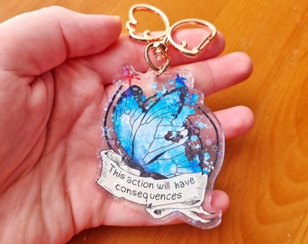 Può includere: Un portachiavi in acrilico trasparente con una farfalla blu decorata con glitter. Il portachiavi ha un anello in metallo dorato e una chiusura argentata. La farfalla ha un design blu e bianco e il testo "This action will have consequences" è scritto su uno stendardo sotto la farfalla.