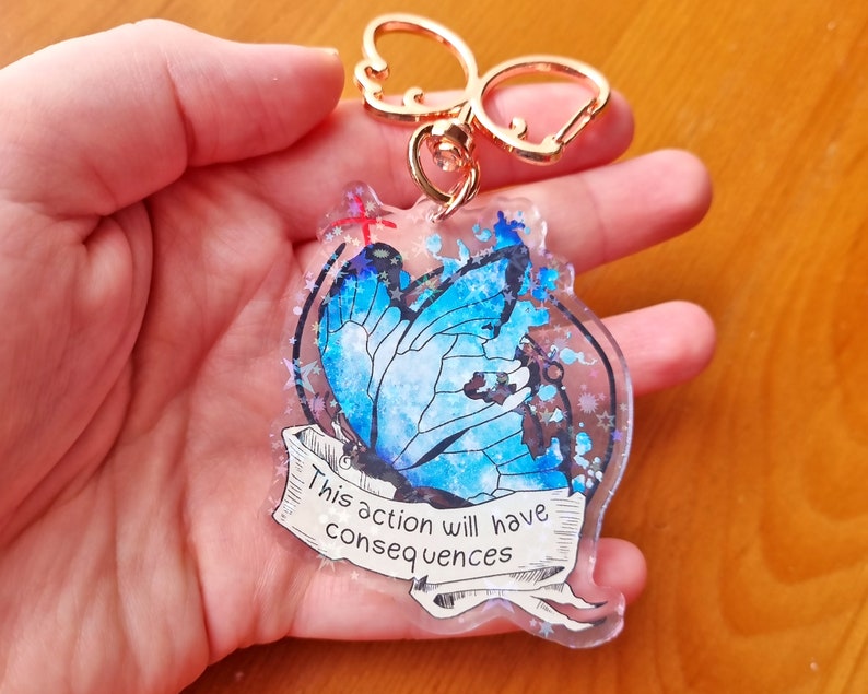 May include: A blue butterfly with glitter accents on a clear acrylic keyring. The keyring has a gold metal ring and a silver clasp. The butterfly has a white and blue design and the text "This action will have consequences" is written on a banner below the butterfly.