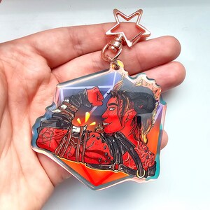 May include: A clear acrylic keyring featuring a red-skinned character with horns, flexing a fist. The character is wearing a black leather-like outfit. The keyring has a star-shaped clasp and a geometric border with iridescent accents.