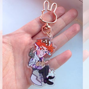 May include: A clear acrylic keyring featuring a rose gold-toned metal clasp shaped like a rabbit head. The charm displays an illustrated character with orange hair, wearing a white coat, holding a brown rabbit, with a white rabbit nearby.