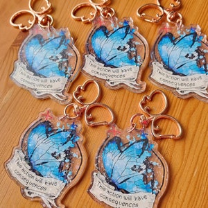 May include: Five blue butterfly charm keyrings with gold-coloured metal rings. Each charm features a blue butterfly with glitter and the text "This action will have consequences".