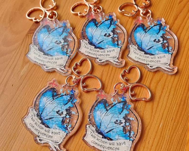 May include: Five blue butterfly charm keyrings with gold-coloured metal rings. Each charm features a blue butterfly with glitter and the text "This action will have consequences".