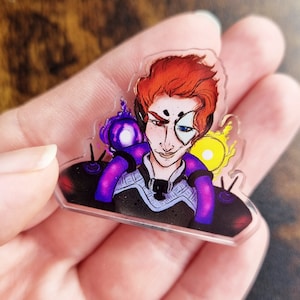May include: A clear acrylic pin featuring a cartoon illustration of a red-haired person with one blue eye and one yellow eye. The person is wearing a black and grey outfit with purple and yellow accents.