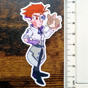 Pu&ograve; includere: Un adesivo fustellato raffigurante un personaggio con capelli arancioni, che indossa un camice bianco e guanti viola, mentre tiene in mano un piccolo coniglio marrone. Un righello accanto mostra un'altezza di circa 19 cm.