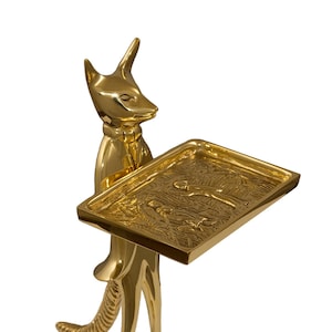 May include: A golden fox figurine holding a rectangular tray. The fox is standing upright, with its head and tail raised. The tray has a detailed relief design. The figurine is made of a shiny, reflective metal.