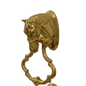 May include: A gold-toned horse head door knocker with a decorative, ornate design. The knocker is attached to a round, gold-toned base.
