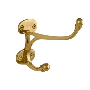 May include: A brass double coat hook with a polished finish. The hook has two curved arms with rounded ends, and is mounted on a decorative backplate with screw holes. The hardware is designed for wall mounting.