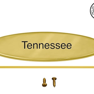 May include: A gold-colored, oval-shaped sign with the word "Tennessee" in black lettering. Includes a matching gold-colored bar and two screws. A quality guarantee seal is in the upper right corner.
