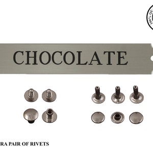 May include: A rectangular, silver-colored sign with the word "CHOCOLATE" in black, accompanied by several silver rivets. The sign has rounded edges and pre-drilled holes for mounting. Includes an extra pair of rivets.