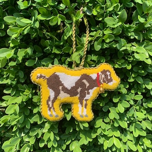 May include: A handmade, yellow goat ornament with brown and white markings, a red collar, and a gold bell. The ornament is made of needlepoint and has a gold rope for hanging. The background is a dense, green bush.