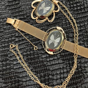 Könnte beinhalten: Ein goldfarbenes Mesh-Armband und eine passende Anhängerkette, beide mit ovalen schwarzen Glaskabochons mit eingraviertem weißem Schmetterlingsmotiv. Das Armband hat ein breites Mesh-Band und einen Verschluss, während die Kette eine feine Gliederkette besitzt.