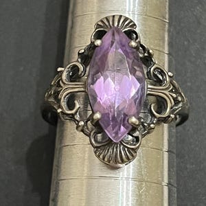 May include: A silver-toned ring featuring a marquise-cut light purple gemstone set in a decorative metal frame with intricate scrollwork and fluted details, positioned on a metal ring mandrel with visible size markings.