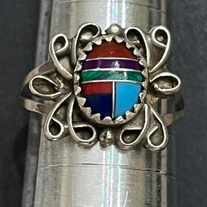 May include: A silver ring featuring a central oval inlay of multi-colored stone segments in red, purple, green, dark blue, and light blue, surrounded by a decorative silver wire scrollwork frame, mounted on a metal ring sizing mandrel.