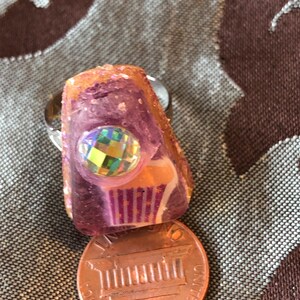 Könnte beinhalten: Ein rosafarbener Harzring mit einem klaren, schillernden Stein und einem Design einer rot-weiß-blauen gestreiften Flagge. Der Ring liegt neben einem US-Penny.
