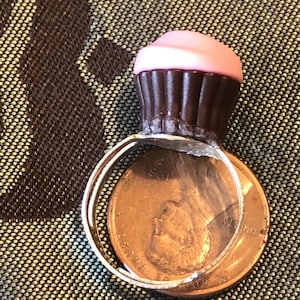Könnte beinhalten: Ein Cupcake-Ring mit rosa Zuckerguss und brauner Cupcake-Basis. Der silberne Ring liegt auf einem Penny.