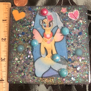 Puede incluir: Una sirena amarilla y azul con alas rosas y un collar de perlas está rodeada de purpurina y cuentas azules y blancas. La sirena está en un marco de resina cuadrado.