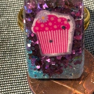 Könnte beinhalten: Ein rechteckiger Harzring mit einer rosa Cupcake-Grafik und einer kleinen Blume darauf, eingebettet in einen klaren Harzblock, der mit violettem und blauem Glitzer gefüllt ist. Der Ring liegt auf einer kupferfarbenen Ein-Cent-Münze.