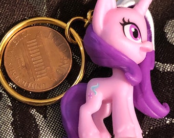 May include: A purple and pink My Little Pony keychain with a gold ring and a penny attached. The pony has a purple mane and tail, a pink body, and a blue and white horn.