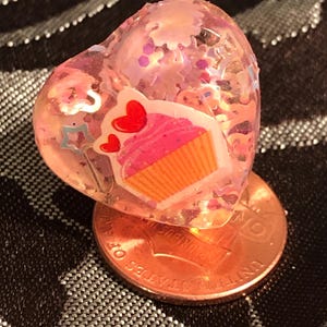 Könnte beinhalten: Ein rosafarbener herzförmiger Harz-Anhänger mit Cupcake-Design und Glitzer im Inneren. Der Anhänger liegt auf einem US-Penny.